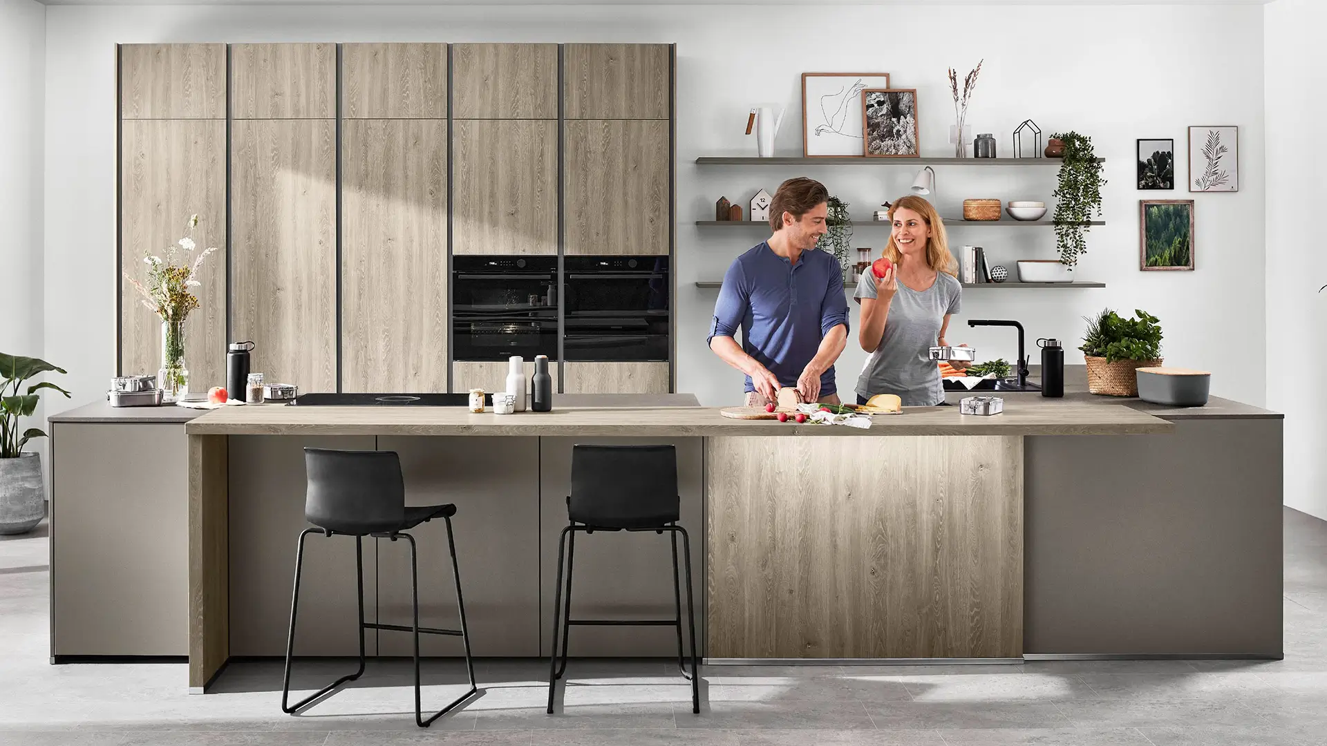 Modern offene Küche mit Holzfronten, Kochinsel und zwei Personen beim Zubereiten von Essen.